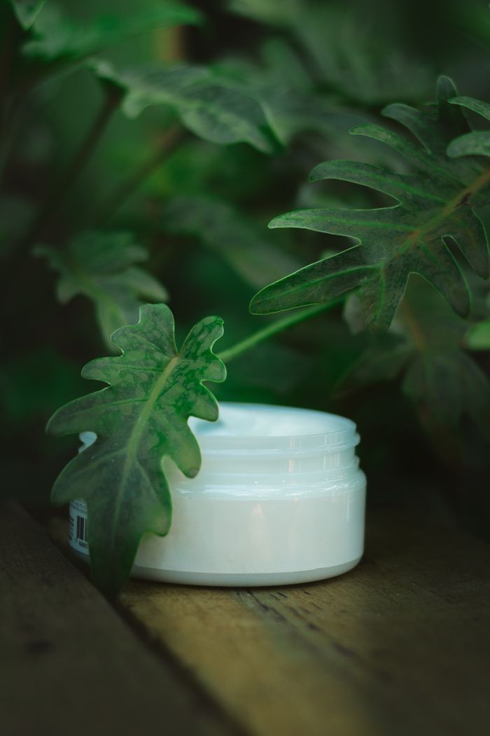 Rutina bio completă de îngrijire a corpului: Exfoliere, hidratare și fermitate Close-up of skincare cream jar amidst lush green leaves, highlighting eco-friendly beauty.
